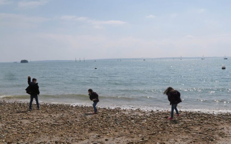 Three children walk and play on a rocky beach near the water’s edge during week 22, with a calm sea and distant boats in the background under a partly cloudy sky.