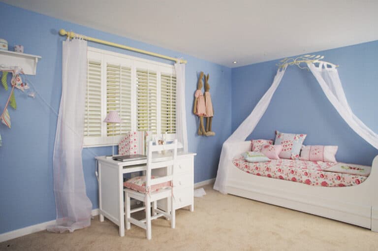 A kids' room with light blue walls, a white desk and chair, and a white daybed with pink patterned bedding and canopy. Sheer window treatments frame the window, while dresses hang on hooks near the bed.