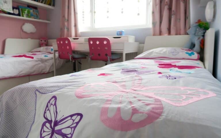 A bright, tidy kids' room features two single beds with butterfly bedding, pink walls, a white desk with two pink chairs, and a window with light curtains letting in natural light—perfect for organisation and cheerful play.