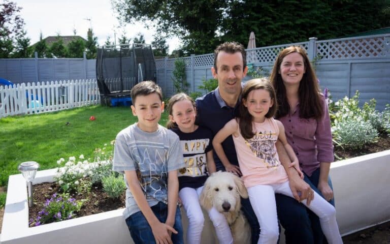 A Me and Mine moment: In June, a family of five with a white dog poses in their back garden. Three children sit in front, parents behind them, all smiling. A trampoline and white fence can be seen in the background.