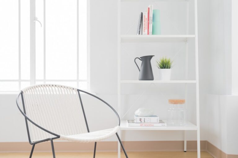 A minimalist home room with a white chair, a white shelf holding books, a black jug, a small potted plant, a glass jar, and magazines. Sunlight streams in through a large window, enhancing the serene minimalist vibe.