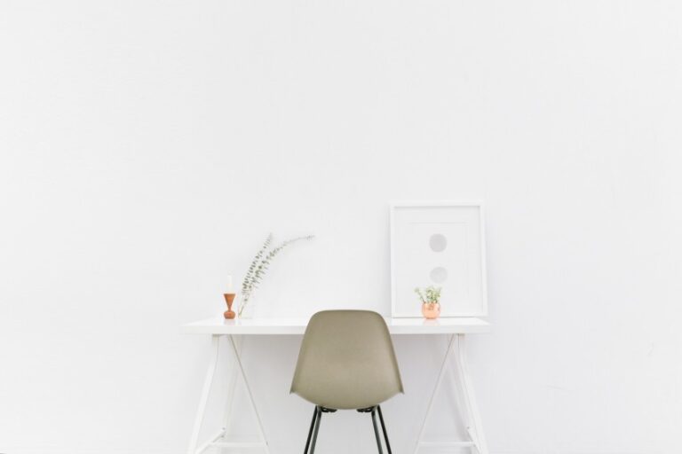 Minimalist home workspace with a grey chair at a white desk against a white wall. The setup features a framed print, two small vases with plants, and a decorative item, creating an open space that welcomes more light.