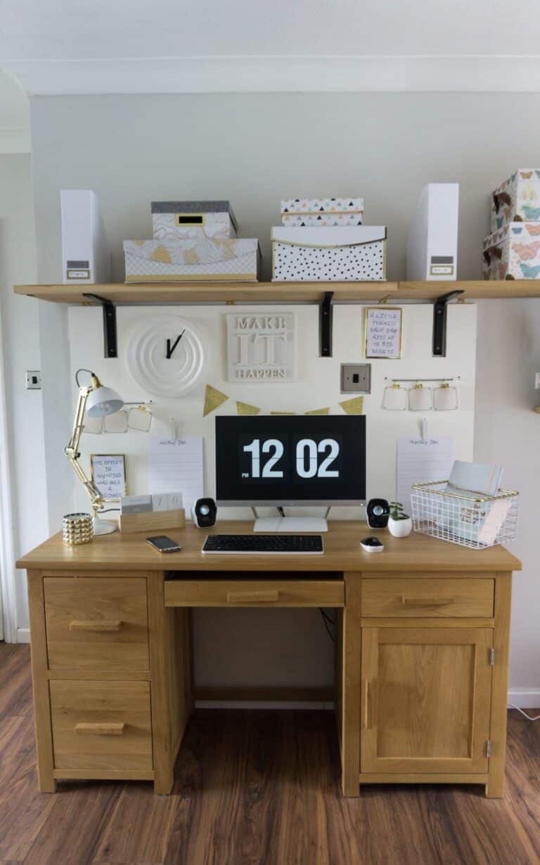 A tidy wooden desk with a computer displaying 12:02, gold lamp, keyboard, and mouse. Above the desk is a shelf with boxes, decor, and stationery. A gold and white noticeboard adds to the workspace’s minimalist, organised feel.