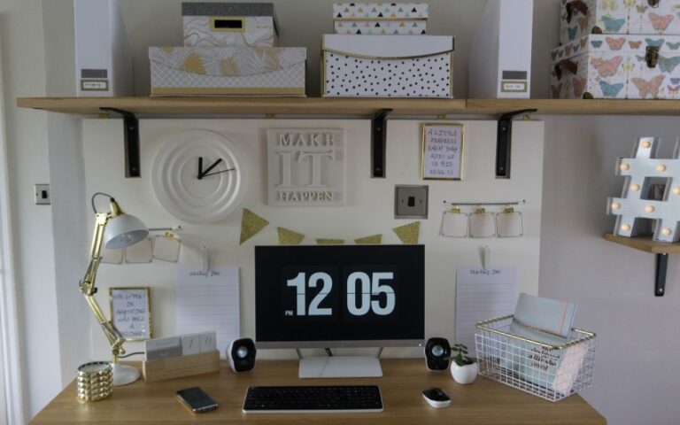 A tidy home office desk with a computer displaying 12:05, surrounded by organised supplies, a gold lamp, boxes, notepads, and decorative wall items—including a Make It Happen sign and a stylish home office noticeboard on the floating shelf above.