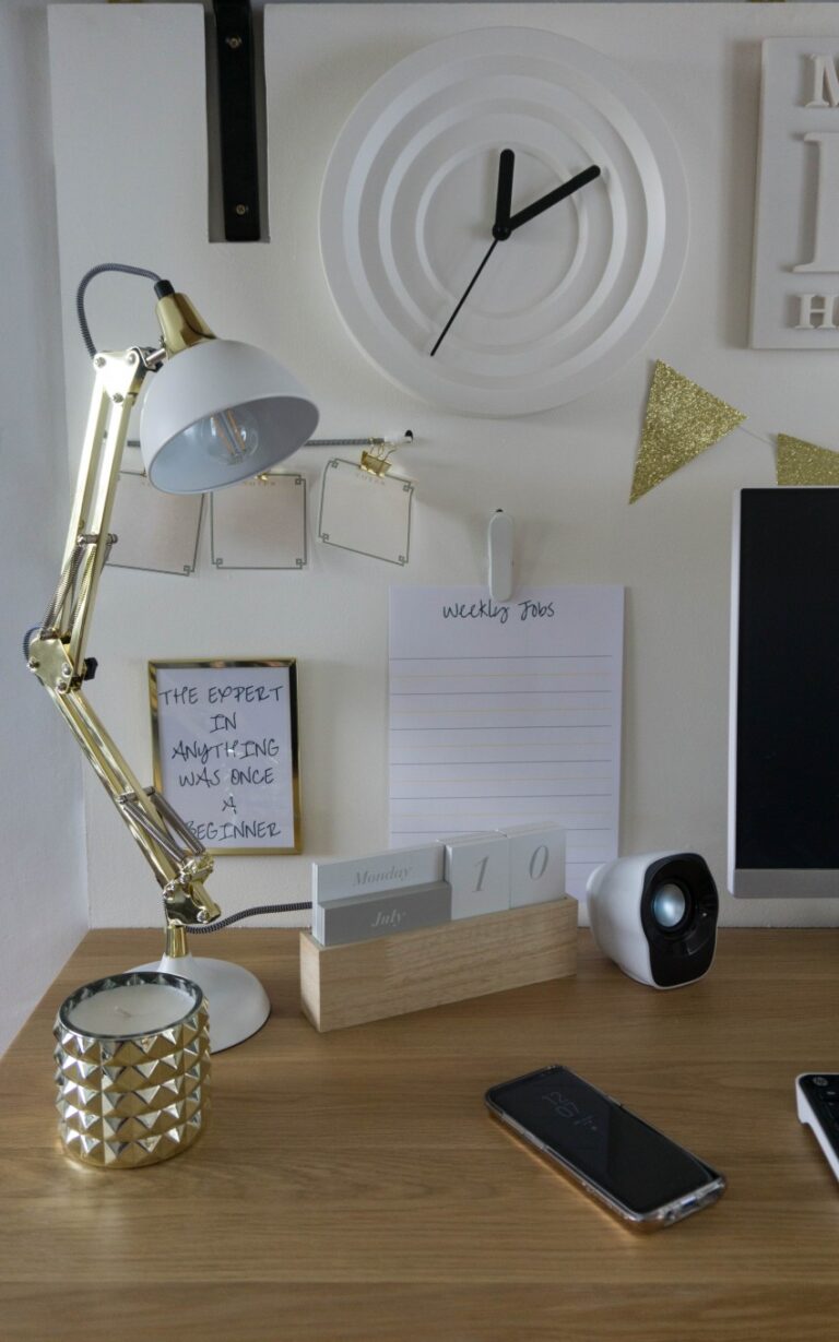 A tidy wooden desk with a lamp, a calendar, a Weekly Jobs notepad, a candle, quote card, mobile phone, and part of a computer monitor. Above hangs bunting, a modern wall clock, and a stylish gold and white noticeboard for your home office.