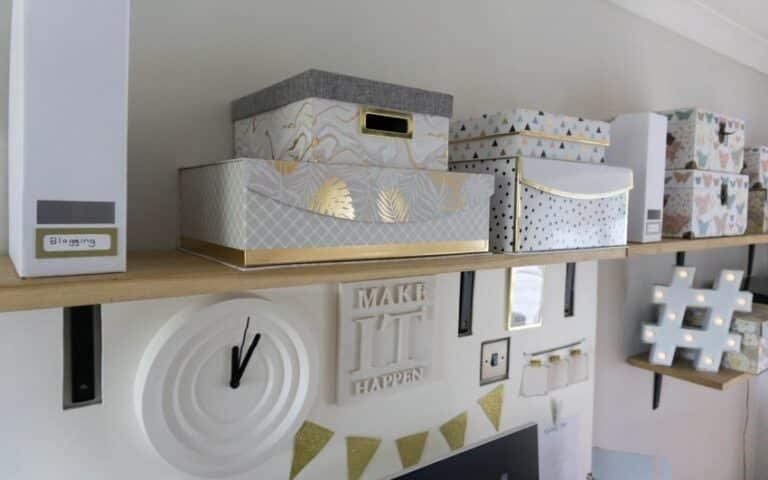 A wooden shelf holds decorative storage boxes with patterns like marble, gold leaves, and polka dots. Below, wall decor features a DIY noticeboard, clock, Make It Happen sign, bunting, and a light-up hashtag symbol.