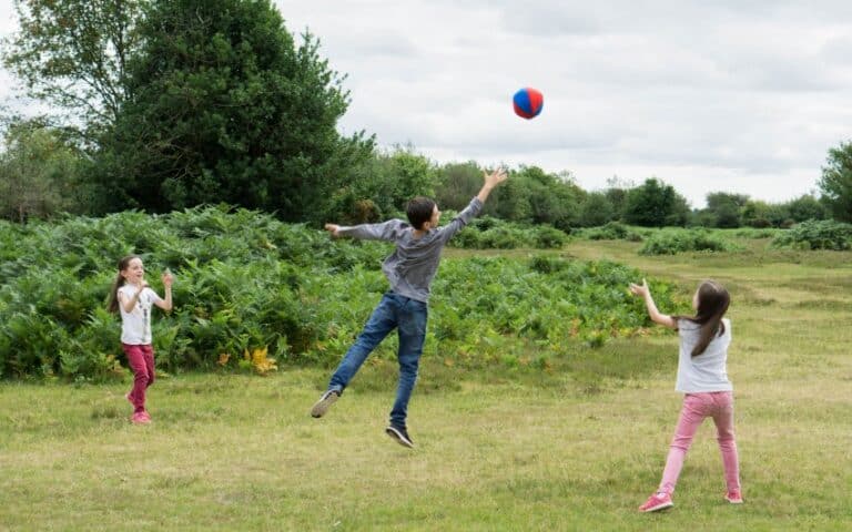 Three children play catch with a red and blue ball in a grassy field, surrounded by greenery under a cloudy sky—a perfect scene for any weekend in pictures. One child leaps to catch the ball whilst the other two watch and wait.