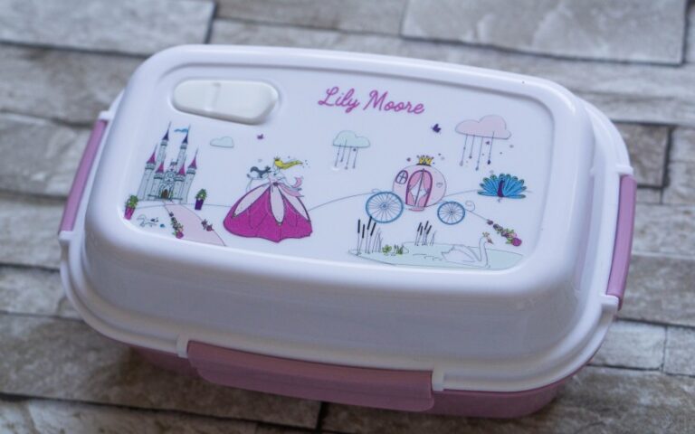 A personalised lunchbox by Petit-Fernand in white and purple, featuring “Lily Moore” on top and decorated with princess-themed illustrations—perfect for packed lunch ideas, pictured on a tiled surface.