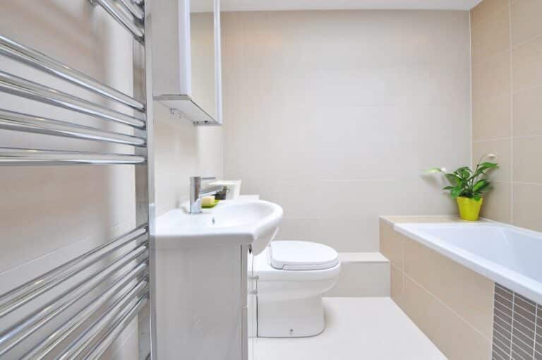 Modern bathroom features white fittings, including a basin, toilet, and bath. A chrome towel rail is on the left, and a small green plant in a yellow pot sits on the ledge by the bath. Light beige tiles complete this stylish family bathroom.