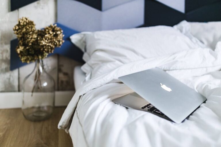 A silver laptop sits partially closed on an unmade bed with white sheets; beside the bed is a clear vase holding dried flowers, adding a touch of home decor to the cosy and relaxed scene.