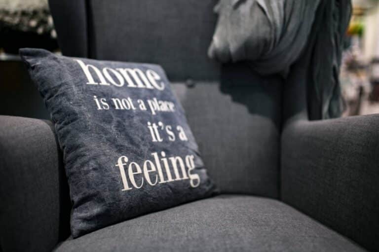 A dark grey armchair with a matching cushion that reads home is not a place its a feeling in white text. A soft grey throw is draped over the chair’s arm—one of those simple ways to be happy and feel happier at home.