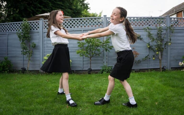 Two young girls in school uniforms and Start-Rite School Shoes hold hands and spin around, smiling and playing on a green lawn in a back garden with a grey wooden fence and plants in the background.