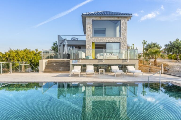 Modern two-storey villa with large windows, blue-tiled roof, and stone accents—perfect for family holidays—featuring a sun terrace with loungers and a swimming pool, set amid trees and plants under a clear sky.