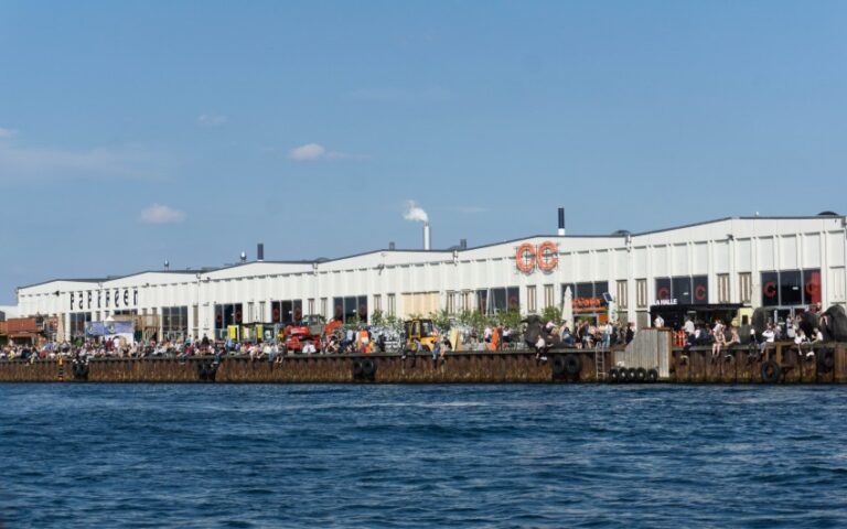 A large white waterfront warehouse with crowds gathered along the pier, under a blue sky and water in the foreground—perfect for a Copenhagen travel adventure or capturing moments during a holiday abroad with children.