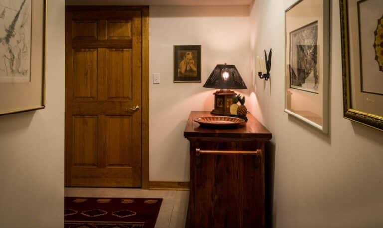 A warmly lit hallway with white walls, wooden door, framed art, and a wooden side table holding a lamp, decorative bowl, and small sculpture offers great hallway decorating ideas for creating a welcoming first impression. A patterned rug is partially visible on the floor.