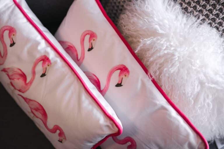 Two white pillows with pink flamingo prints and pink piping—featuring fashionable motifs—are next to a fluffy white cushion on a textured grey surface.