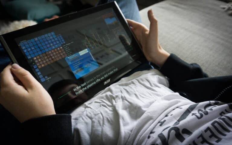 A person holding an Acer tablet on their lap, playing a video game with inventory and menu screens visible. The scene highlights the importance of Online Safety while relaxing indoors, as they wear a white T-shirt and a dark jacket.
