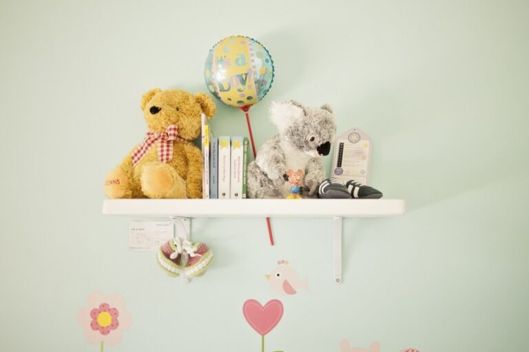 A white wall shelf holds a yellow teddy bear, a grey koala plush, children's books, a birthday balloon, tiny shoes, and toys—perfect touches for preparing for baby. The light green wall features pink flower and bird decals below the shelf.