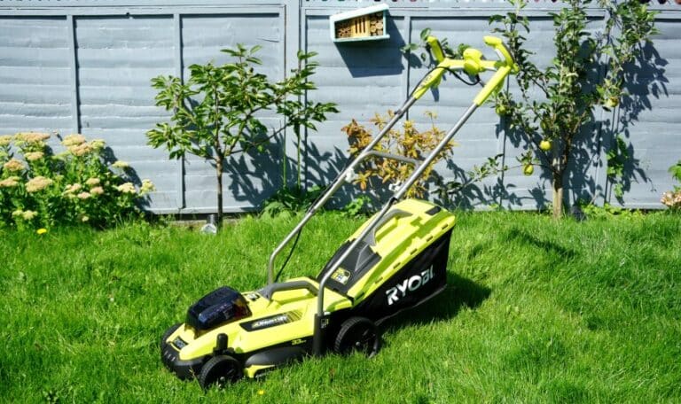 A bright green Ryobi Cordless Lawnmower sits on lush grass in a sunny garden, positioned near a grey wooden fence with plants and a small bird box attached to it.