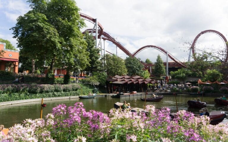 A roller coaster winds through a lush, green amusement park—one of my favourite places to travel—with boats floating on a pond and blooming pink flowers in the foreground under a partly cloudy sky.