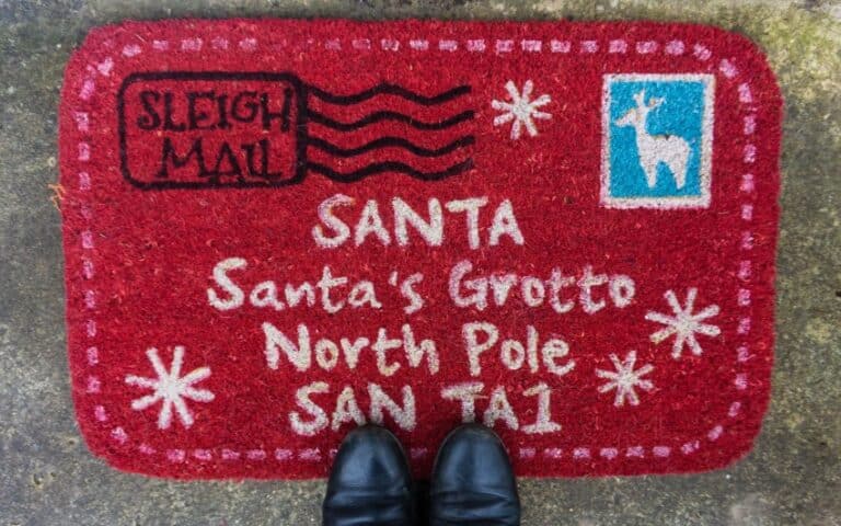 A red doormat designed like a letter, with white text reading Father Christmas, Father Christmas’ Grotto, North Pole, SAN TA1, postage stamp, and SLEIGH MAIL mark. Perfect for a Christmas Front Door to offer a festive Christmas Welcome. Two black shoes are visible at the bottom.