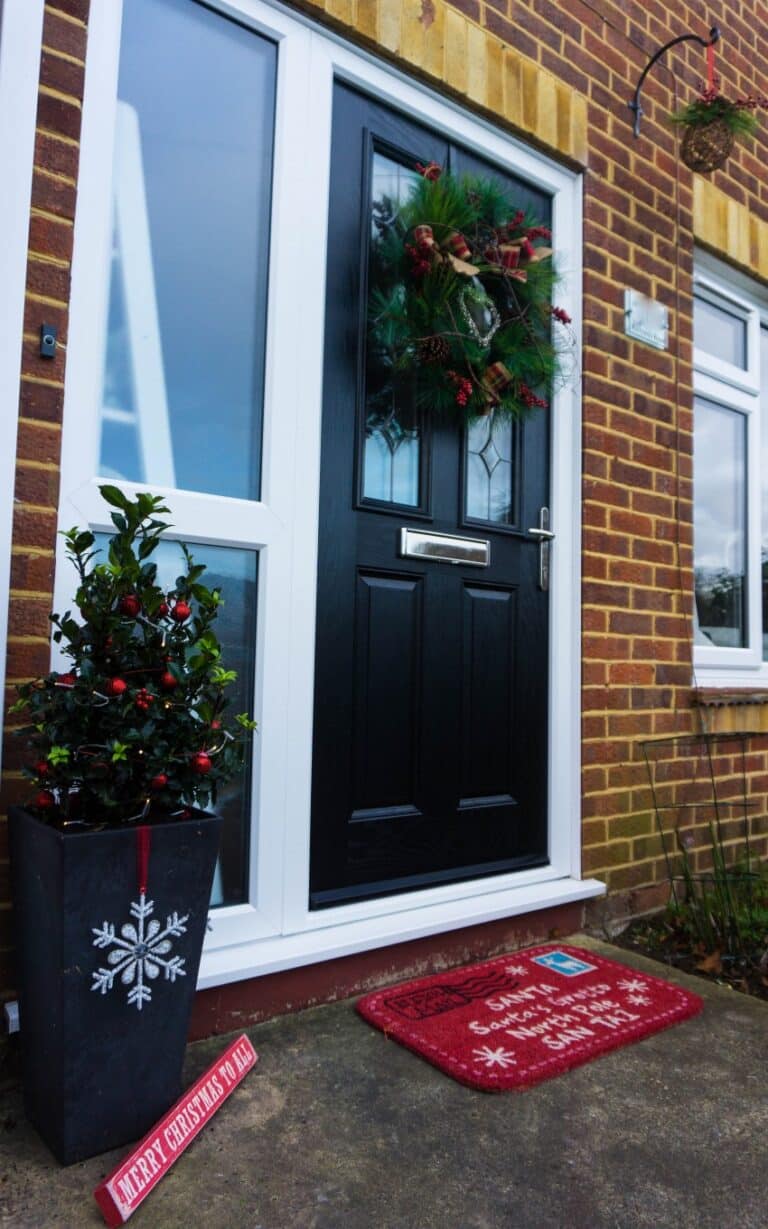 A festive Christmas front door with a cheerful wreath stands beside a white-framed window. A red doormat with holiday designs, a snowflake pot holding a plant, and a Merry Christmas Ya Filthy Animal sign complete this charming Christmas welcome.