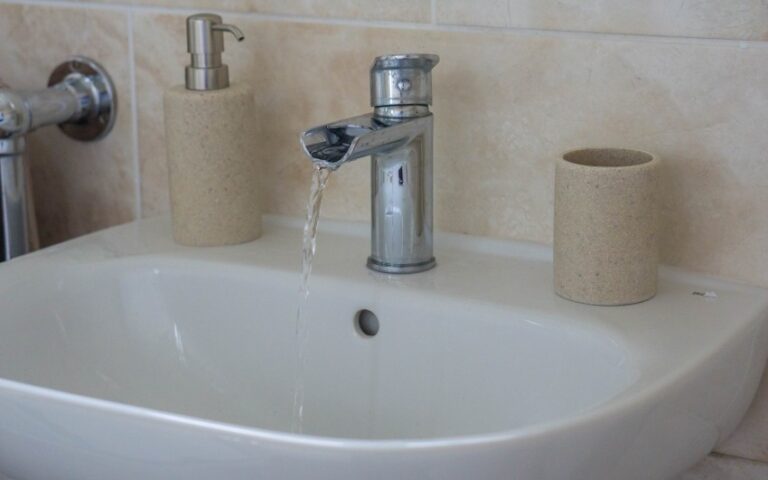 A bathroom basin with water running from a modern silver tap. Styled To Sell, the beige soap dispenser and toothbrush holder sit neatly on either side of the basin against a tiled wall background.