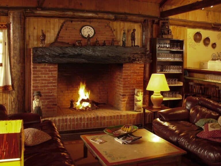 A cosy living room with a brick fireplace lit, perfect for winter home preparation, surrounded by wooden walls, leather settees, a coffee table with snacks, a lamp, and shelves with glasses. Small figurines and a clock sit on the mantelpiece.