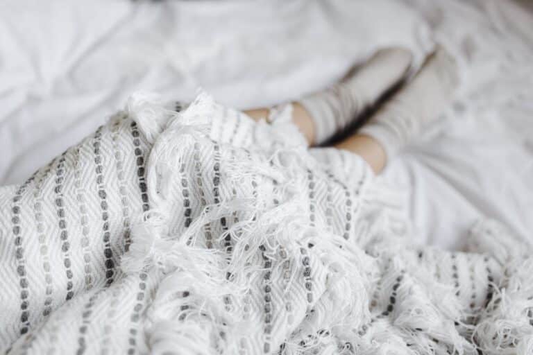 A person wearing light-coloured socks is lying on a bed, partially covered with a white blanket featuring black stripes and fringed edges—a perfect scene to cosy up and unwind in a cosy home.