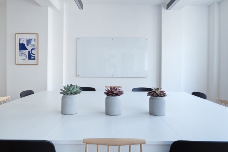 A modern, minimalist conference room designed for home efficiency, featuring a large white table with three potted succulents, black and wooden chairs, a blank whiteboard on the wall, and abstract art framed on the left.