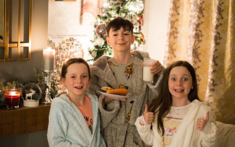 Three smiling children in pyjamas stand in a cosy living room decorated for Christmas. One boy holds biscuits and milk, while the girls give a thumbs up by the lit tree—capturing the joy of Christmas 2017 in heartwarming Christmas photos.