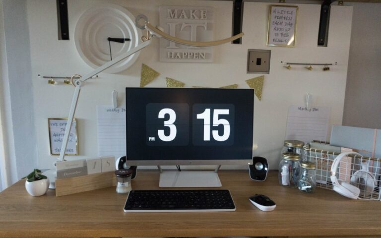 A tidy desk setup with a monitor displaying 3:15 pm, white keyboard and mouse, desk lamp, speakers, jars, headphones, a small plant, and decorative wall art and notes about planning permission above the desk.
