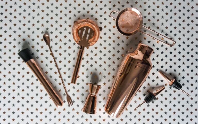 A set of shiny copper cocktail tools, perfect for crafting tasty cocktails, including a shaker, strainer, jigger, spoon, tongs, pourers, and a brush—arranged on a white surface with small gold star patterns.