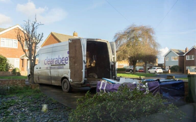 A white Clearabee van with open rear doors is parked on a residential drive, offering an efficient rubbish removal service. Several pieces of large debris are piled beside the van, with houses and trees visible in the background.