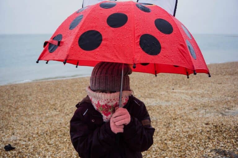 A person stands on a pebbly beach holding a red umbrella with black spots, resembling a ladybird. Bundled in a knitted hat and scarf, they face the cold, rainy day—capturing the mood for this week 46 weekend in pictures vlog.