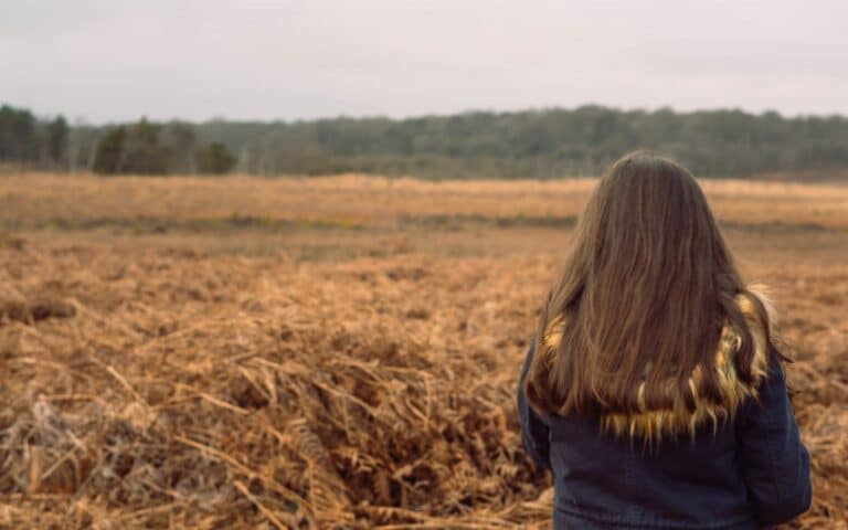 A person with long brown hair and a dark jacket stands facing a wide, dry field with tall grass, capturing the quiet mood of Week 47’s weekend in pictures, gazing towards a distant tree line under an overcast sky.