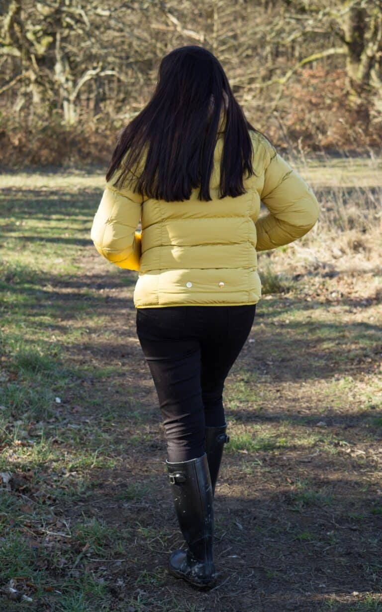 A brave person with long dark hair, wearing a mustard yellow puffer jacket, black trousers, and black boots, walks along a grassy sunlit path surrounded by trees. The person is seen from behind.
