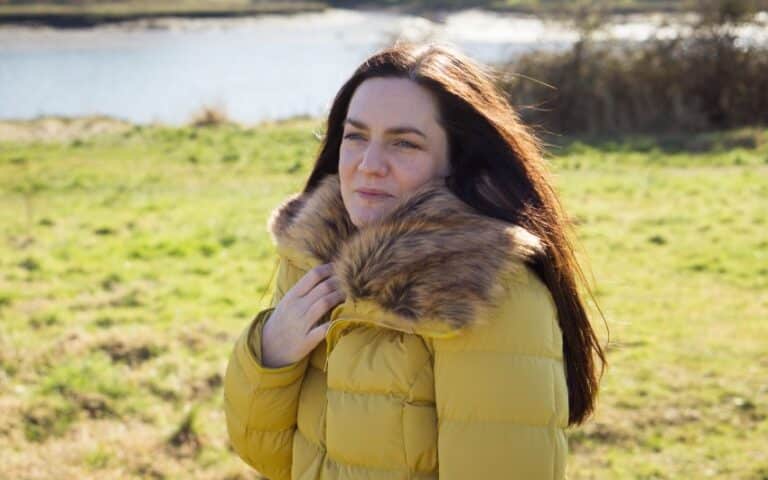 A brave woman with long brown hair stands outdoors on a sunny day, wearing a mustard yellow coat with a large, furry collar. Green grass and a body of water are visible in the background.