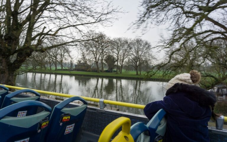 A person in a winter coat and hat sits on the upper deck of an Original Tour bus, looking at a calm river and leafless trees in Windsor on a cloudy day.