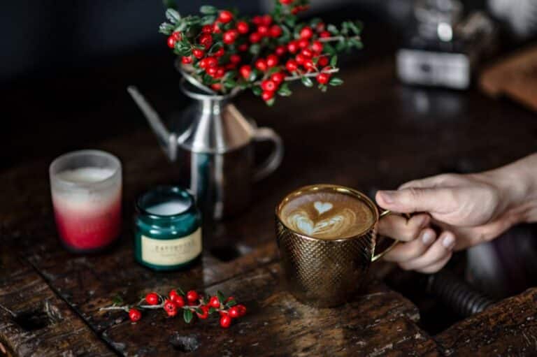 A hand holds a gold cup of coffee with heart-shaped latte art on a rustic wooden table, surrounded by red berries, a candle, a green jar, and a silver jug with berry branches that add to the cosy home fragrance.