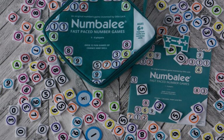 Numerous colourful numbered tokens, perfect for kids' maths, are scattered on a wooden surface around a teal Numblee game pouch and instruction booklet for ages 6+, advertising fast-paced number games.