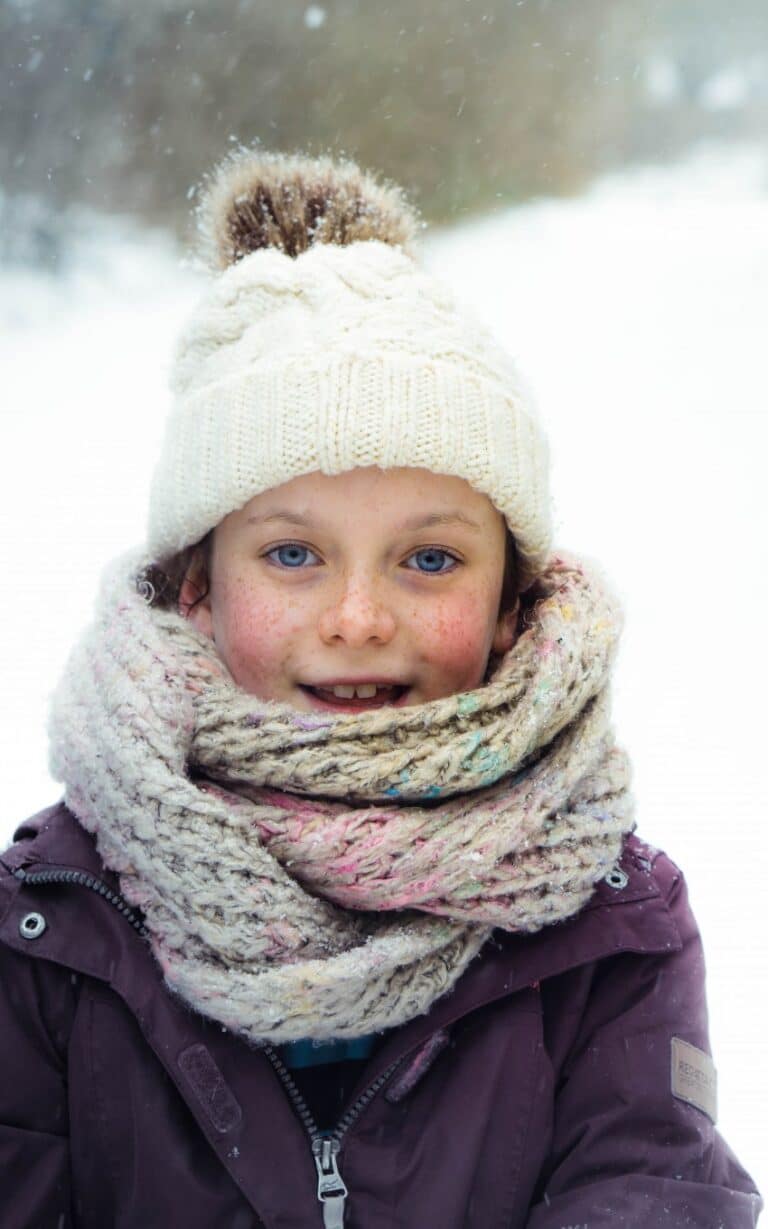 A smiling child wearing a white knitted hat, a thick knitted scarf, and a purple winter coat stands outside in the snow, capturing the magic of Snow Days as snowflakes fall around them—perfect for a cosy 2018 vlog moment.