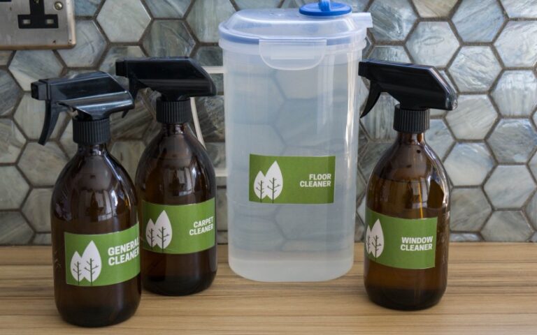 Four labelled cleaning bottles sit on a wooden counter: general cleaner, carpet cleaner, window cleaner (in amber spray bottles), and floor cleaner (in a clear plastic container). These natural cleaning products are perfect for home cleaning. A tiled splashback is in the background.