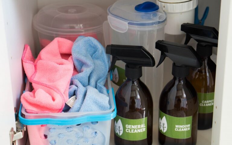 Spray bottles labelled general cleaner, window cleaner, and carpet cleaner sit in a cupboard beside natural cleaning products and a blue basket of pink and blue microfibre cleaning cloths for home cleaning.