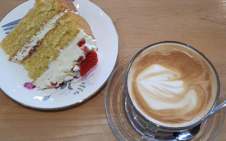 A slice of sponge cake with cream and strawberries on a floral plate next to a glass cup of cappuccino with latte art, both on a wooden table—just one sweet moment from my Weekend In Pictures, Week 56.