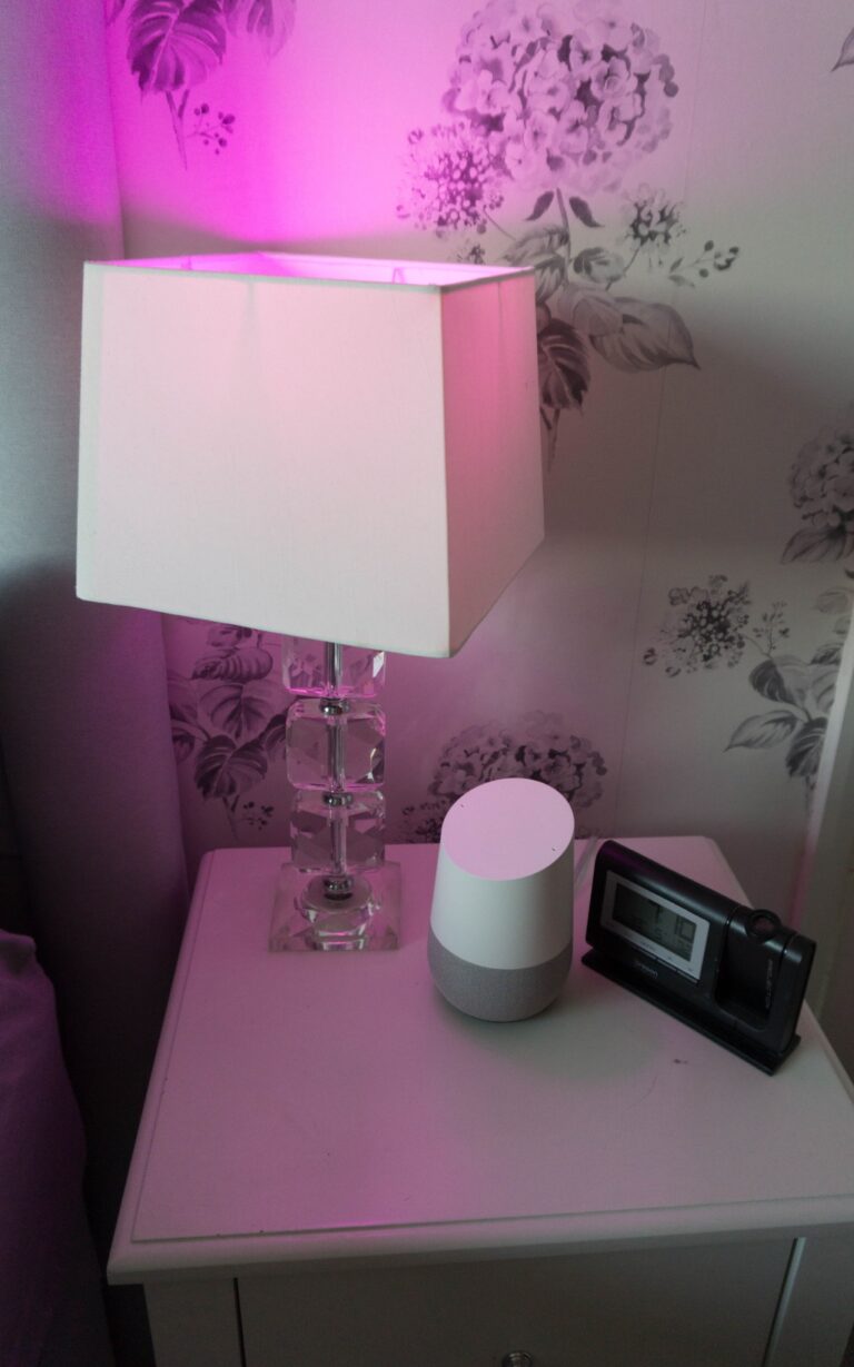 A bedside table with a crystal lamp glowing with pink light, a white smart speaker, and a digital alarm clock sits against floral-patterned wallpaper—perfect for any smart home tour.