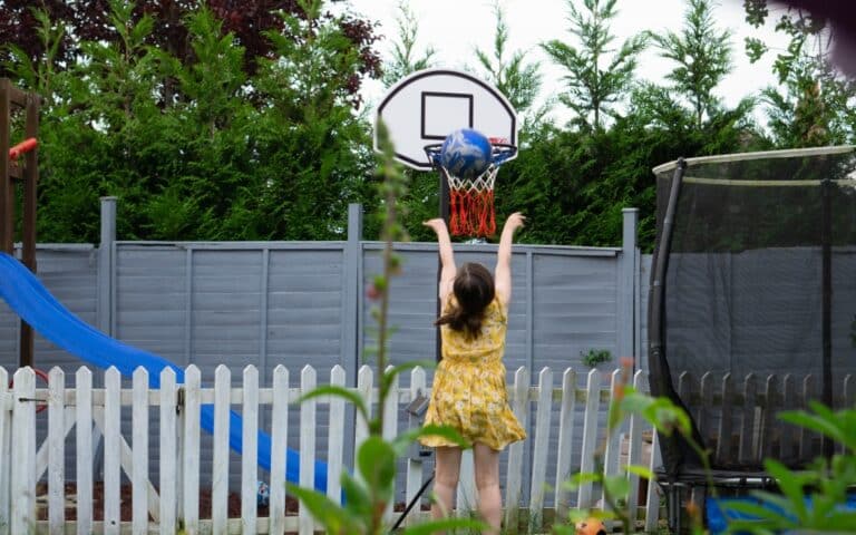 A girl in a yellow dress jumps to shoot a basketball into a hoop in the back garden, surrounded by a white picket fence, grey fence, blue slide, trampoline, and greenery—capturing a fun Weekend In Pictures moment.