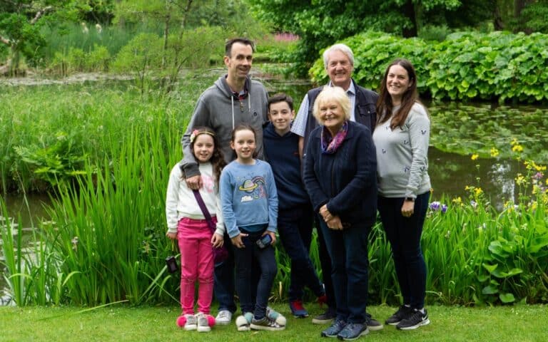 A family of seven, including three children and four adults, stand together smiling in a lush green garden with tall grass, yellow flowers, and a pond, capturing a joyful Fathers’ Day for Weekend In Pictures.