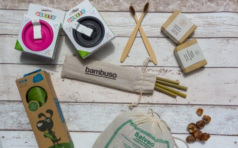 Flat lay for Week 61: eco-friendly products on a wooden surface, featuring reusable cups, bamboo toothbrushes, soap bars, metal straws in a cloth bag, a green bottle in a box, and soap nuts—perfect for your Weekend In Pictures.