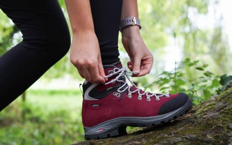 A person in black leggings ties the laces of a red walking boot whilst standing on a mossy log outdoors, with green foliage blurred in the background—showcasing their love of walking boots.